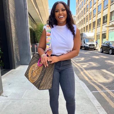 She is wearing awhite top and holding a handbag on her right shoulder.